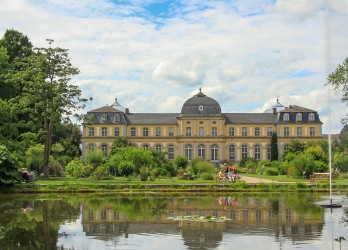 Das Bonner Schloss mit Schlossgarten. Im Vordergrund ein Gewässer, in dem sich das Schloss spiegelt.