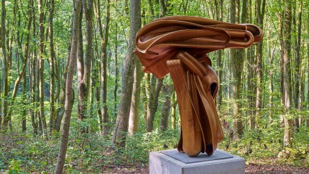 Eine abstrakte, bronzefarbene Skulptur auf einem Steinsockel im Wald.