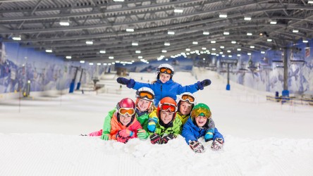 Eine Gruppe von Kindern im Schnee – mit der großen Piste der Skihalle im Hintergrund.