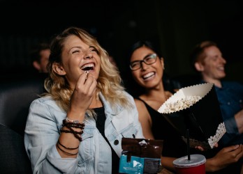 Zwei Frauen sitzen lachend im Kino und essen Popcorn.