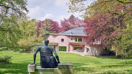 Eine sitzende Skulptur von hinten fotografiert. Im Hintergrund eine Villa mit rosa Fassade.